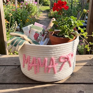 Op de afbeelding: Een witte touwmand met het woord "MAMA" in roze garen. De mand bevat tuinhandschoenen, zaadzakjes, een kleine troffel en een geranium in pot met rode bloemen. De mand staat op een houten oppervlak.