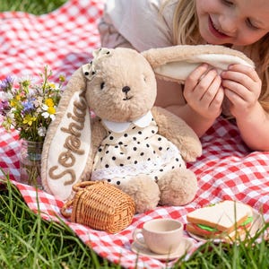 May include: A plush tan rabbit toy with long ears, wearing a cream-colored dress with black polka dots and a white collar. The name Sophia is embroidered in brown on one ear. The rabbit has a small cream bow on its head and soft fur texture.