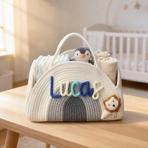 May include: A white and gray woven diaper caddy with a rainbow design and the name "Lucas" in blue and teal letters. The caddy contains diapers, rolled blankets, and a penguin plushie. A lion applique is on the side. The caddy sits on a wooden table.