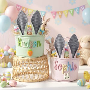Könnte beinhalten: Zwei Osterkörbe mit Hasenohren, einer hellgrün und einer rosa, jeweils mit einem Namen auf der Vorderseite gestickt. Der grüne Korb trägt den Namen "Hudson" und hat eine Karottenapplikation. Der rosa Korb trägt den Namen "Sophie" und hat eine Hasenapplikation. Ostereier und Stoffhasen sind darin.