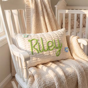 May include: A cream-colored pillow with the name "Riley" embroidered in green yarn, adorned with small floral accents. The pillow rests on a white rocking chair, draped with a cream-colored knitted blanket. A crib with teddy bears is visible in the background.