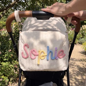 May include: Cream-colored stroller bag with the name "Sophie" in pastel-colored knitted letters. The bag is attached to a stroller with wooden handles. The background shows greenery and sunlight.