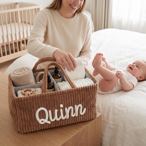 Puede incluir: Una cesta de almacenamiento de pana marrón con dos asas y el nombre Quinn bordado en letra blanca en la parte frontal. El interior está dividido en secciones que contienen una manta beige, juguetes de madera, frascos y pañales blancos.