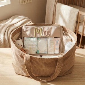 May include: A tan corduroy tote bag with a clear zippered pouch labeled "Mia" containing baby essentials. Inside the tote are diapers, wipes, lotion, a bottle, a rolled towel, and a stuffed animal. The bag has two handles and is on a wooden surface.
