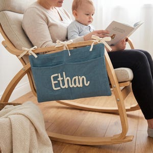May include: A blue fabric storage organizer with two pockets attached to the arm of a wooden rocking chair. The organizer features the name Ethan in white raised lettering and is secured to the chair frame with cream-colored fabric ties.