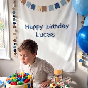 K&ouml;nnte beinhalten: Ein wei&szlig;er Geburtstagshintergrund mit blauen und beigen Wimpeln, kreisf&ouml;rmigen Papiergirlanden und dem blauen Schriftzug "Happy birthday Lucas". Ein mit bunten Bausteinen und Kerzen verzierter Kuchen steht vor dem Banner mit blauen Luftballons.