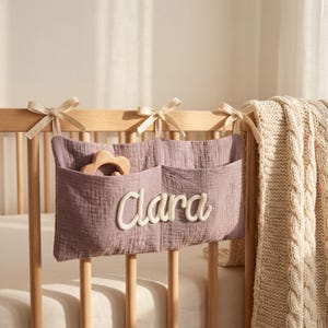 May include: A dusty purple fabric hanging organizer with two pockets, attached to a wooden crib rail with three cream-colored ribbon ties. The name Clara is embroidered in white script on the front, and a wooden flower toy sits in one pocket.