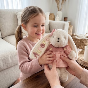 May include: A plush cream-colored rabbit toy wearing a pink and white gingham dress with a matching bow on its ear. The rabbit has the name Aurora embroidered in multi-colored yarn on one of its long, floppy ears, featuring a soft, fuzzy texture.