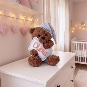 Könnte beinhalten: Ein brauner Plüschteddybär mit einer hellblau-weiß karierten Schlafmütze mit dem Text Goodnight, der ein passendes hellblau-weiß kariertes Kissen hält, auf dem der Name Olivia in einer hellrosa Schreibschrift gestickt ist.