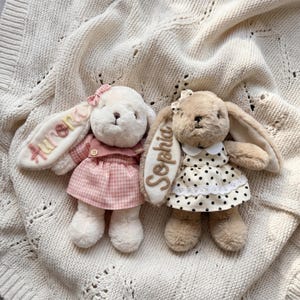 May include: Two plush rabbit toys with long ears. The left rabbit is cream-colored, wearing a pink gingham dress and a small bow, with the name Aurora embroidered on its ear. The right rabbit is tan, wearing a white dress with black polka dots and a lace trim, with the name Sophia embroidered on its ear.