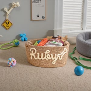May include: A woven dog toy basket with the name "Ruby" in cream-colored letters and a bone accent. The basket contains dog toys and treats. A blue ball, a green leash, and other dog toys are scattered around the basket. The background includes a dog bed and wall decor.