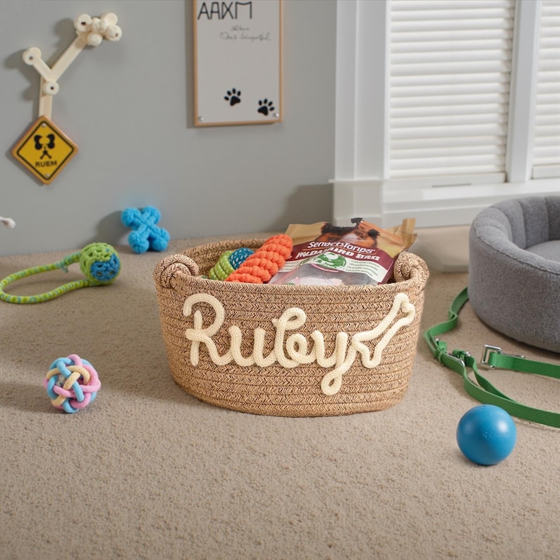 May include: A woven dog toy basket with the name "Ruby" in cream-colored letters and a bone accent. The basket contains dog toys and treats. A blue ball, a green leash, and other dog toys are scattered around the basket. The background includes a dog bed and wall decor.