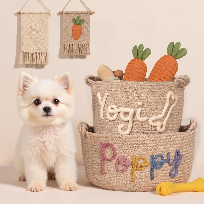 May include: A small, fluffy white dog sits next to two woven baskets. The top basket has the name "Yogi" and the bottom basket has the name "Poppy". The baskets are filled with dog toys, including two orange carrot-shaped toys. Two decorative wall hangings are in the background.