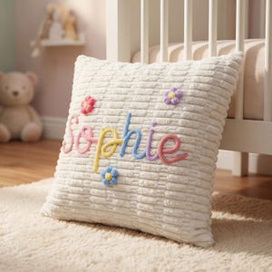 May include: Cream-colored corduroy pillow embroidered with the name "Sophie" in colorful thread. The pillow features small, embroidered flowers in pink, purple, and blue. The pillow is on a cream-colored rug.