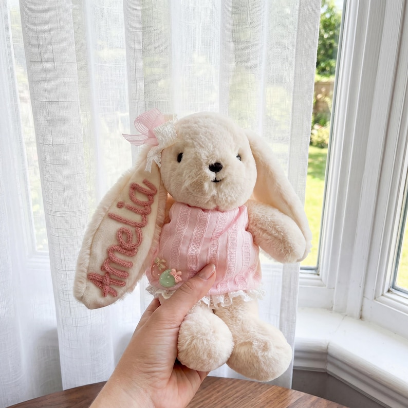 May include: A cream-colored plush rabbit toy wearing a light pink ribbed sweater with a lace trim. The rabbit has a pink bow on its left ear, and the name Amelia is embroidered in pink thread on the inner side of the same ear.