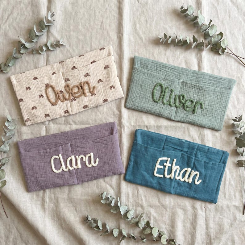 May include: Four rectangular fabric organizers with embroidered names. One is cream with brown rainbows and the name Owen, one is sage green with Oliver, one is dusty purple with Clara, and one is teal blue with Ethan. Each has a front pocket.
