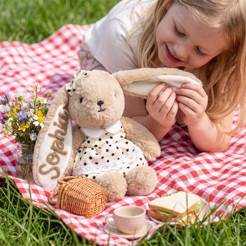 May include: A plush tan rabbit toy with long ears, wearing a cream-colored dress with black polka dots and a white collar. The name Sophia is embroidered in brown on one ear. The rabbit has a small cream bow on its head and soft fur texture.