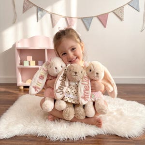 May include: Three plush stuffed bunnies with long floppy ears. Each bunny features a name embroidered on one ear: Aurora in pink, Sophia in brown, and Amelia in pink. The bunnies wear small outfits including a pink gingham dress and a polka dot dress.