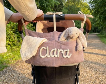 May include: A mauve fabric stroller organizer featuring the name Clara embroidered in white script. The organizer is attached to a brown leather-look stroller handle with cream ribbons and contains a white muslin cloth and a plush beige bunny toy.
