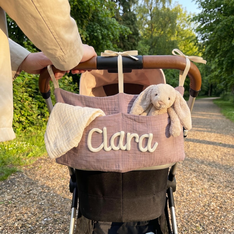 May include: A mauve fabric stroller organizer featuring the name Clara embroidered in white script. The organizer is attached to a brown leather-look stroller handle with cream ribbons and contains a white muslin cloth and a plush beige bunny toy.
