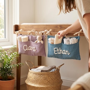 May include: Two rectangular fabric storage pockets in purple and blue, tied to a wooden frame with cream ribbons. The purple pocket features the name Clara in white script, and the blue pocket features the name Ethan in white script.