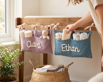 May include: Two rectangular fabric storage pockets in purple and blue, tied to a wooden frame with cream ribbons. The purple pocket features the name Clara in white script, and the blue pocket features the name Ethan in white script.