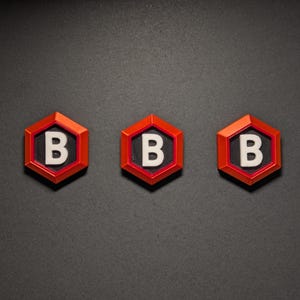 Könnte beinhalten: Drei rot-schwarze sechseckige Objekte mit einem weißen Buchstaben "B" in der Mitte. Sie sind horizontal auf einem dunkelgrauen Hintergrund angeordnet. Die Objekte haben eine glänzende Oberfläche.