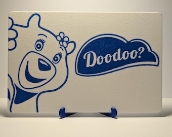 May include: A rectangular white sign with a blue cartoon bear illustration and the word "Doodoo?" in a blue cloud-shaped speech bubble. The sign is propped up on a blue stand.