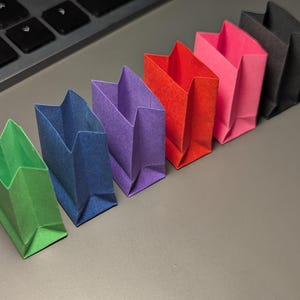 May include: A row of six miniature paper bags in vibrant colors including green, blue, purple, red, pink, and black. Each bag features a rectangular base, folded sides, and an open top, arranged in a diagonal line on a smooth surface.