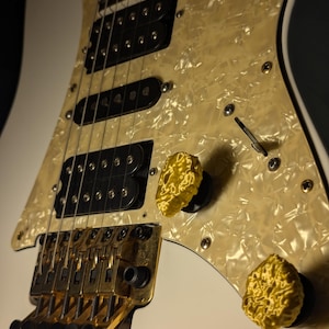 May include: A white electric guitar with a pearlescent pickguard and gold hardware. The guitar features black pickups, gold control knobs, and a tremolo bridge. The body is white, and the pickguard has a textured, off-white appearance.