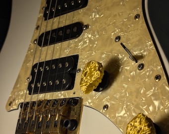 May include: A white electric guitar with a pearlescent pickguard and gold hardware. The guitar features black pickups, gold control knobs, and a tremolo bridge. The body is white, and the pickguard has a textured, off-white appearance.