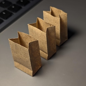 May include: Three small, brown paper bags are arranged in a row, each with a folded top. The bags are made of a textured, kraft paper material. The background is a gray surface, with a portion of a black keyboard visible in the upper left corner.