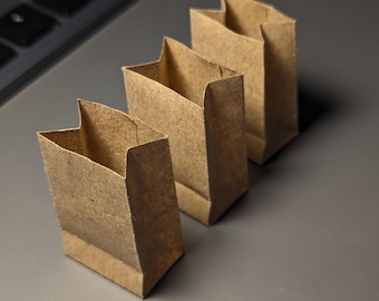 May include: Three small, brown paper bags are arranged in a row, each with a folded top. The bags are made of a textured, kraft paper material. The background is a gray surface, with a portion of a black keyboard visible in the upper left corner.