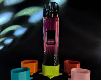 May include: A pink and black vape device with the word "NOVO" printed on it, surrounded by four colored stands. The stands are teal, orange, red, and pink. The device is standing on a lime green stand. The background is black with light patterns.