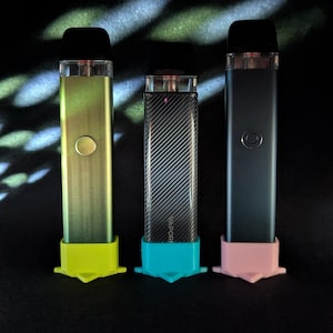 May include: Three vape pens in a row, each with a different color. The first is a light green with a yellow base, the second is a carbon fiber design with a blue base, and the third is a dark gray with a pink base. All have black mouthpieces.
