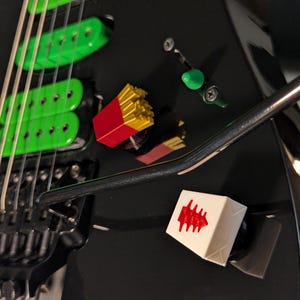 May include: Black electric guitar with green pickups and silver strings. A red and yellow french fry box and a white cube with a red design are placed on the guitar. A green tuning peg is also visible.