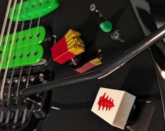May include: Black electric guitar with green pickups and silver strings. A red and yellow french fry box and a white cube with a red design are placed on the guitar. A green tuning peg is also visible.