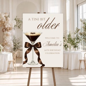 May include: A sign on an easel with the text "A Tini Bit Older" and "Welcome to Amelia's 30th Birthday Celebration." The sign features a martini glass with coffee beans and a brown ribbon. The background shows a party setting with balloons.