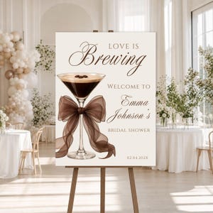Puede incluir: Un letrero rectangular sobre un caballete de madera con el texto Love is Brewing, Welcome to Emma Johnson's Bridal Shower y la fecha 02.04.202X. El diseño incluye una copa de martini llena de café oscuro y un lazo de organza marrón.