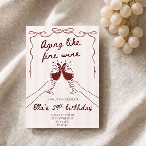 May include: A rectangular birthday invitation card with a light pink striped background. The text reads Aging like fine wine, JOIN US TO CELEBRATE, Ella's 29th birthday, and event details. It features a dark red illustration of two wine glasses clinking.