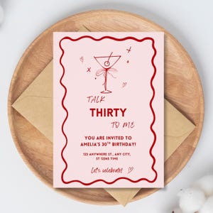 以下が含まれることがあります： 赤い縁とテキストが入ったピンク色の誕生日招待状。「Talk Thirty To Me」と書かれており、アメリアの30歳の誕生日のお祝いの詳細が含まれています。招待状は、茶色の封筒が入った木製のトレイにあります。
