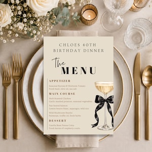 May include: A table setting with a menu for "Chloes 40th Birthday Dinner." The menu lists appetizers, main courses, and dessert options. Gold-colored cutlery and a champagne glass with a black bow are visible. White flowers and candles add to the elegant ambiance.