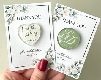 May include: Two personalized pins on white cards with floral borders. One heart-shaped clear pin features the letters J and E with a branch graphic and the date 4.14.26. The other circular sage green pin displays the letters E and D with 4.24.26.