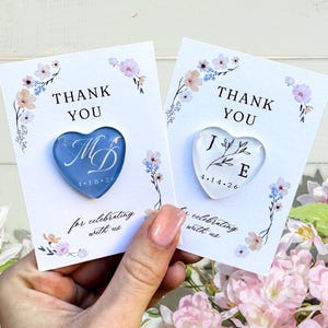 May include: Two heart-shaped magnets on cards that say "THANK YOU". One magnet is blue with "MD" and the date. The other is clear with "JE" and the date. Floral accents and the phrase "for celebrating with us" are also present.