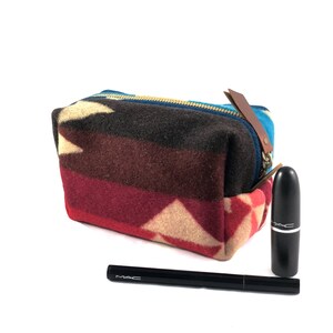 Könnte beinhalten: Eine Kosmetiktasche aus Stoff mit rotem, braunem, blauem und weißem Muster, mit Reißverschluss und braunem Leder-Zugband. Die Tasche liegt auf einer weißen Oberfläche neben einem schwarzen Lippenstift und einem schwarzen Eyeliner-Stift mit dem Aufdruck "MAC".