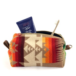 May include: A rectangular toiletry bag made of fleece with a geometric pattern in shades of orange, red, brown, and cream. The bag features a brass zipper, brown leather side tabs, and contains a blue tube of Facial Fuel and a wooden toothbrush.