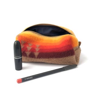 May include: A fuzzy, zippered pouch featuring a horizontal gradient pattern in shades of orange, red, and brown with a geometric triangle design. Beside the pouch sits a black lipstick tube and a black lip liner pencil with a coral-colored tip.