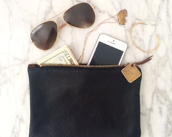 May include: A black leather zippered pouch with a gold zipper and a gold tag that says "Wanderlust". Inside the pouch are a twenty dollar bill, a cell phone, and a pair of aviator sunglasses.