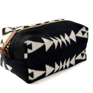 May include: A rectangular zippered pouch made of black and white felted fabric featuring a repeating geometric triangle pattern. The bag includes a gold-toned metal zipper closure with a brown leather pull tab attached to the side of the zipper.