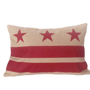 May include: A rectangular pillow with a beige burlap fabric cover featuring the red and white colors of the District of Columbia flag. Three red stars are centered on the top of the pillow.